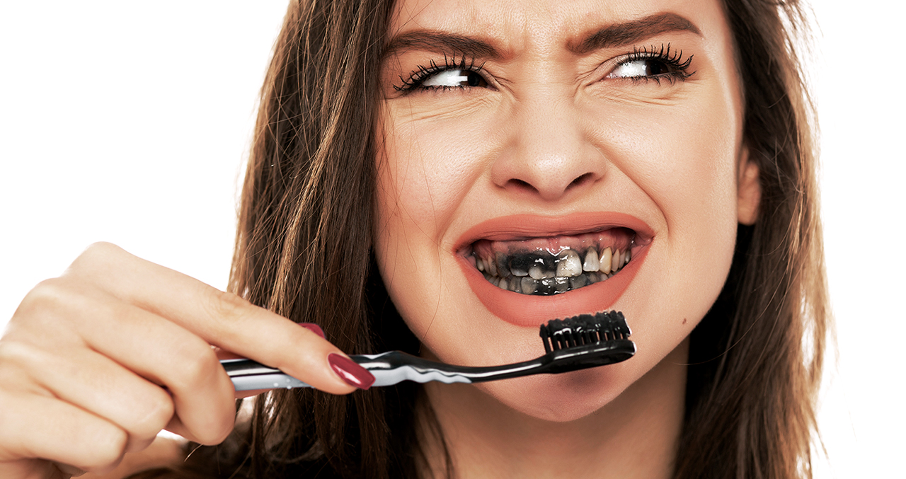 Portrait: Frau putzt sich die Zähne mit schwarzer Aktivkohle und schaut angewidert – ihre Zähne sind ebenfalls schwarz.