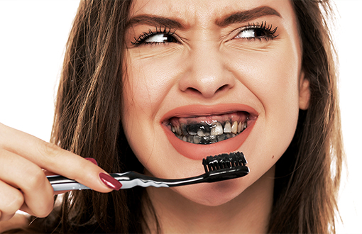 Portrait: Frau putzt sich die Zähne mit schwarzer Aktivkohle und schaut angewidert – ihre Zähne sind ebenfalls schwarz.