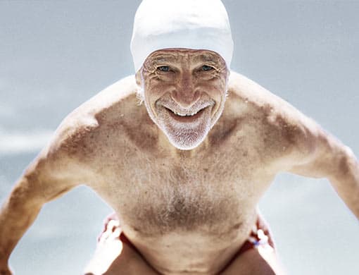 Ein älterer Herr mit weißem Bart, Badekappe und Badehose macht sich bereit zum Startsprung.